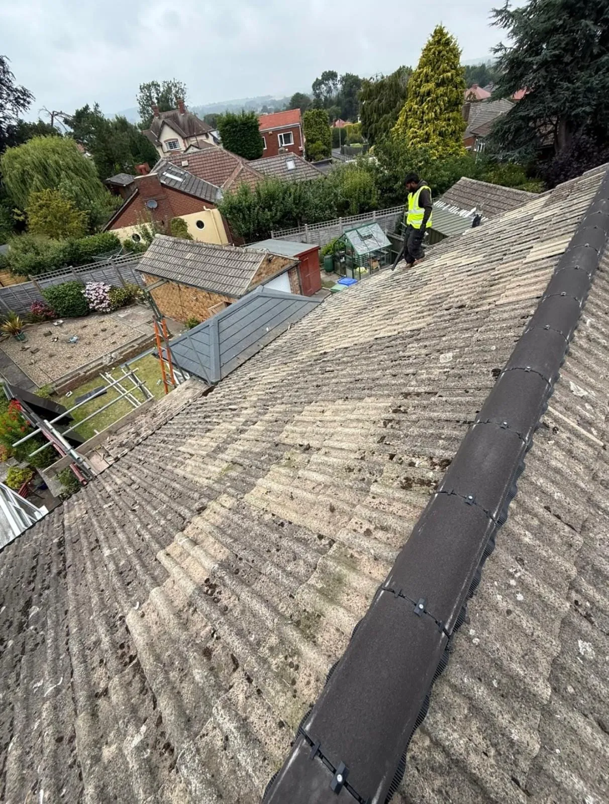 Grey roof slope with a dark ridge, with roofers working above a housing estate