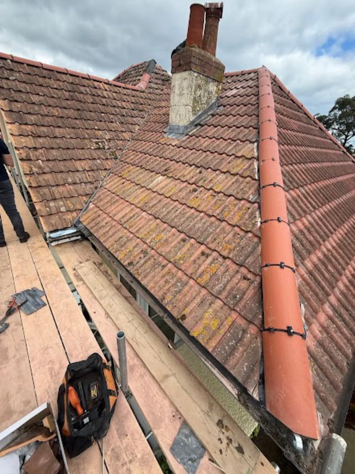 New ridge tiles beside a chimney on a tiled roof, with a roofer’s tool bag on the scaffold
